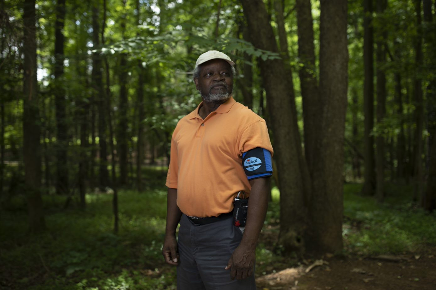 A man in a wooded area looking off camera, wearing a SunTech blood pressure monitor on his left arm